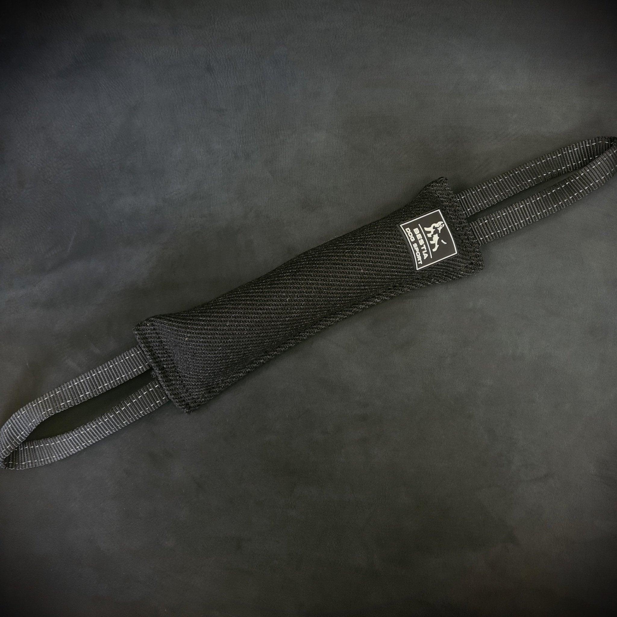 Das BESTIA DOG SPORT – Beißkissen für Hunde aus schwarzem Nylon mit verstärkten Nähten, zwei Schlaufen und Markenlabel, ideal für Hundetraining sowie Biss- und Grifftraining, liegt auf einer dunklen Oberfläche.